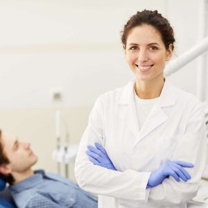 Waist up portrait of female dentist smiling at camera while consulting patient in background, copy space