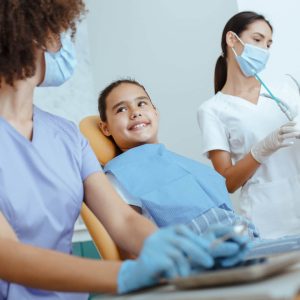 Work of modern dentist orthodontist with children. Millennial friendly african american doctor and european nurse in uniforms, protective masks and rubber gloves start examining little girl, cropped